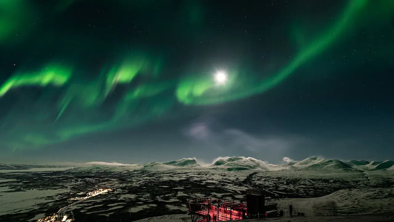 Norrsken över ett snöigt landskap med en upplyst byggnad. Kanske en vy från ett tåg till Abisko.