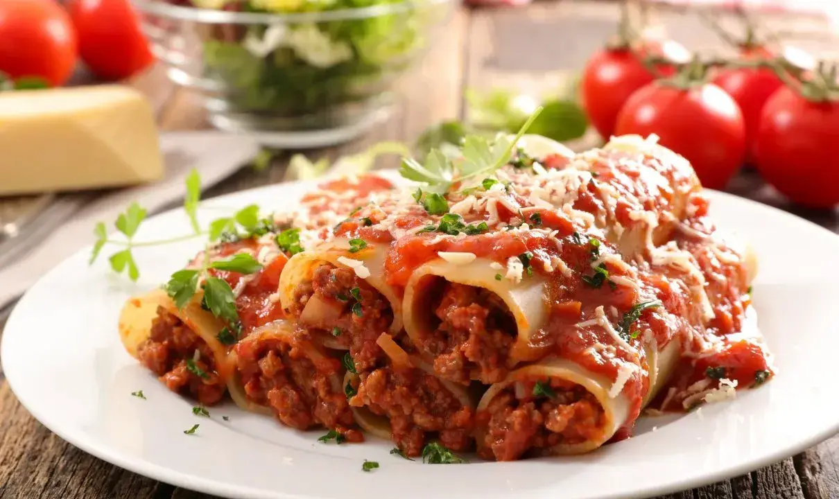 Pyszne cannelloni z mięsem mielonym, polane sosem pomidorowym i posypane parmezanem, z dodatkiem świeżej natki pietruszki.