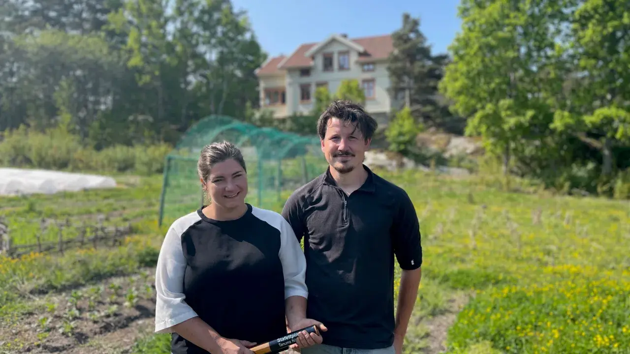 Två personer står på en åker vid Peters place Grebbestad. Bakom dem syns ett hus och grönskande träd.