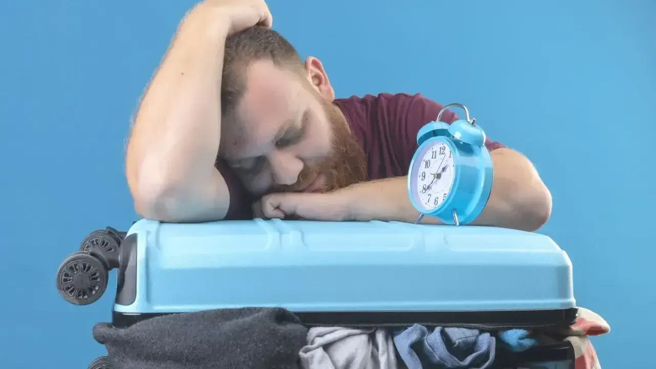 Zmęczony podróżnik śpi na walizce, budzik obok sygnalizuje jetlag.
