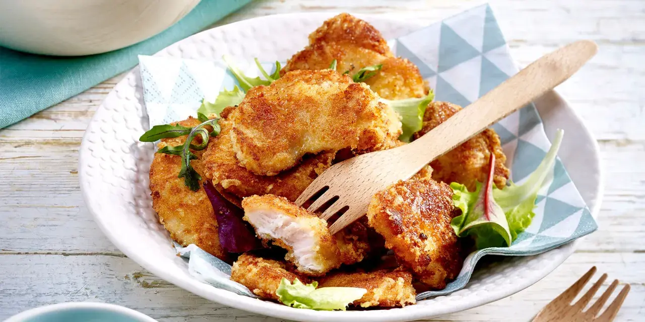 Knusprige Hähnchen-Nuggets, perfekt im Butterschmalz gebraten, auf einem Bett aus Salat. Ein Holzgabel liegt bereit.