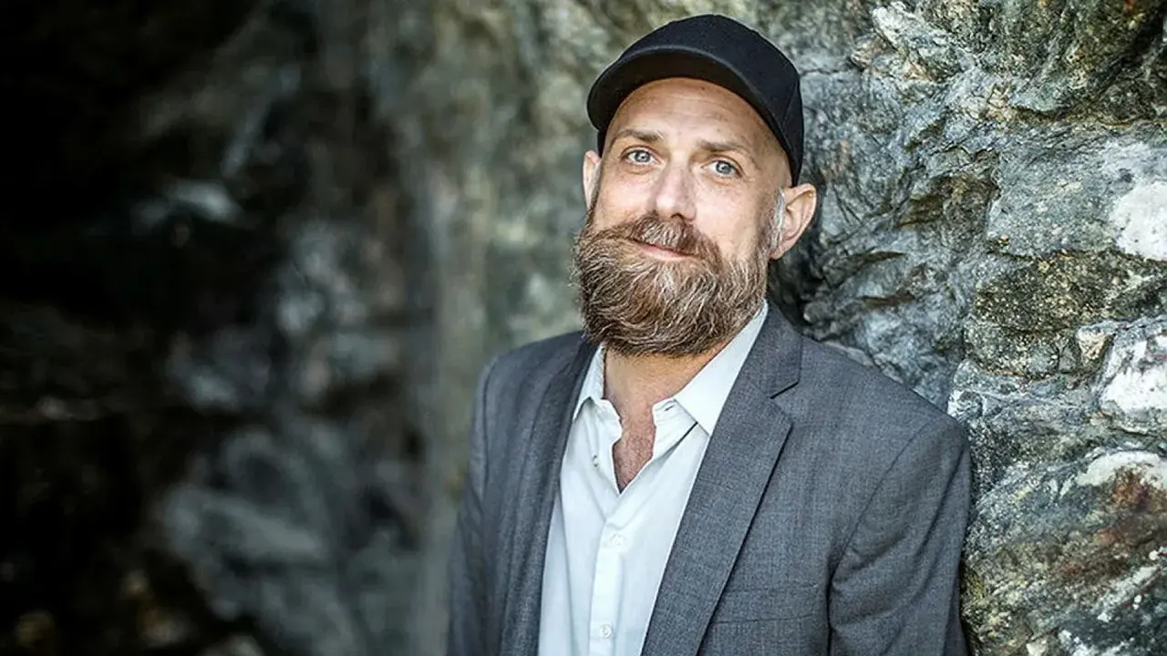 Man med skägg och keps, i grå kavaj, står mot en stenvägg. Kanske en del av en förintelsen serie på SVT.