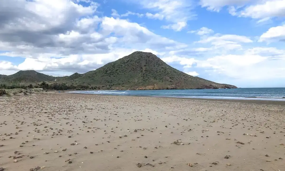 Weite Sandfläche am Playa de los Genoveses mit sanften Wellen und einem grünen Berg im Hintergrund unter einem bewölkten Himmel.
