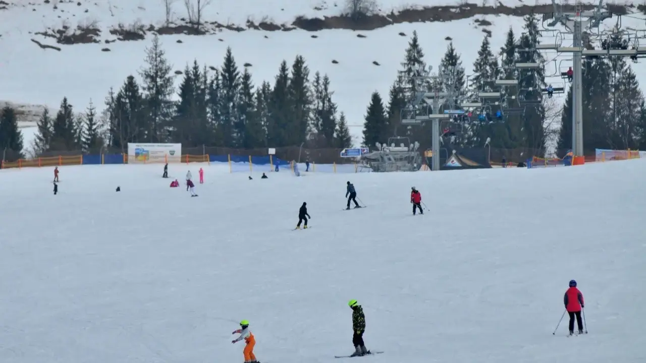 Narciarze i snowboardziści szusują po ośnieżonym stoku w Bukowinie Tatrzańskiej. W tle widać wyciąg krzesełkowy i las.