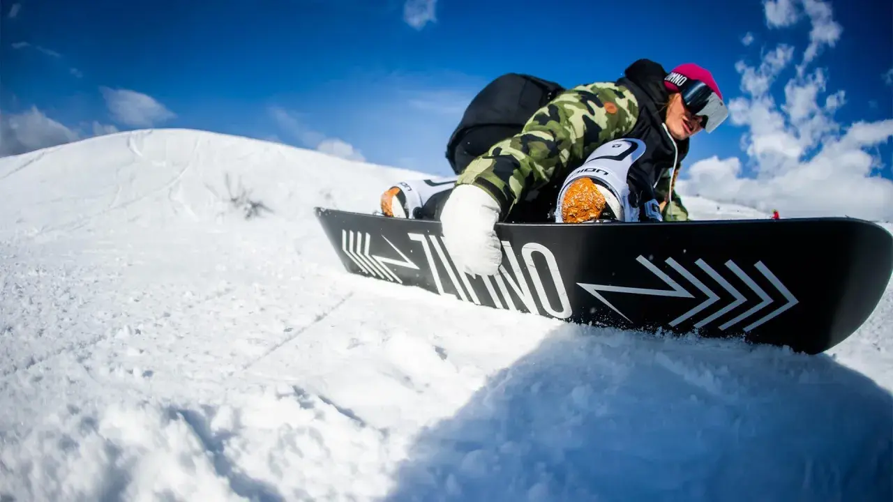Snowboardzista w kamizelce moro i różowej czapce, zjazd na desce. Warto wiedzieć, jak dobrać buty snowboardowe, by czuć się pewnie na stoku.