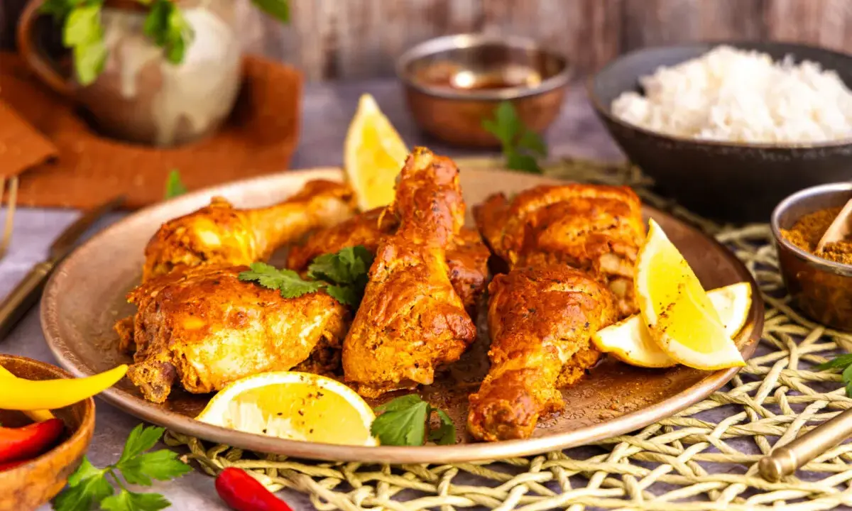 Saftiges Chicken Tandoori auf einem Teller mit Zitronenspalten und Koriander. Daneben Reis und Gewürze.