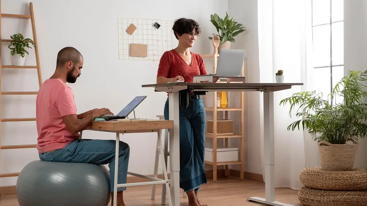 Un homme travaille sur son ordinateur, assis sur un ballon de gym, tandis qu'une femme travaille debout à un bureau réglable. Un espace de travail moderne et dynamique.