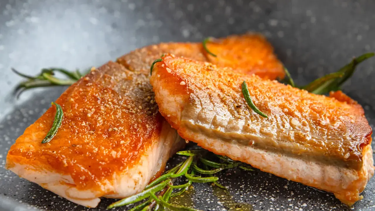 Zwei perfekt gebratene Lachsfilets mit knuspriger Haut und Rosmarin in einer Pfanne.