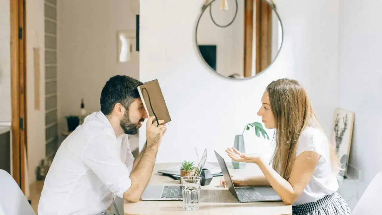 Hombre con personalidad narcisista, frustrado, se cubre la cara con un libro mientras una mujer le habla con exasperación.