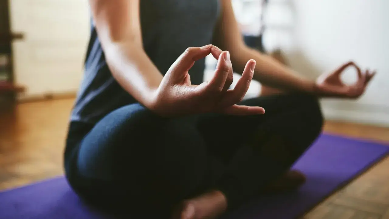 En person sitter i meditationsställning på en lila matta. Händerna vilar i knät med tumme och pekfinger sammanfogade, en vanlig gest för att visa hur man mediterar.
