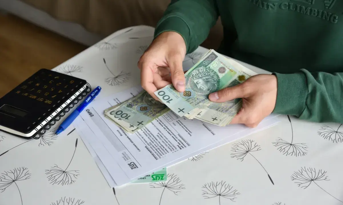 Ręce liczą banknoty 100 zł nad dokumentami ZUS. Nowe zasady zasiłków przedemerytalnych w praktyce.
