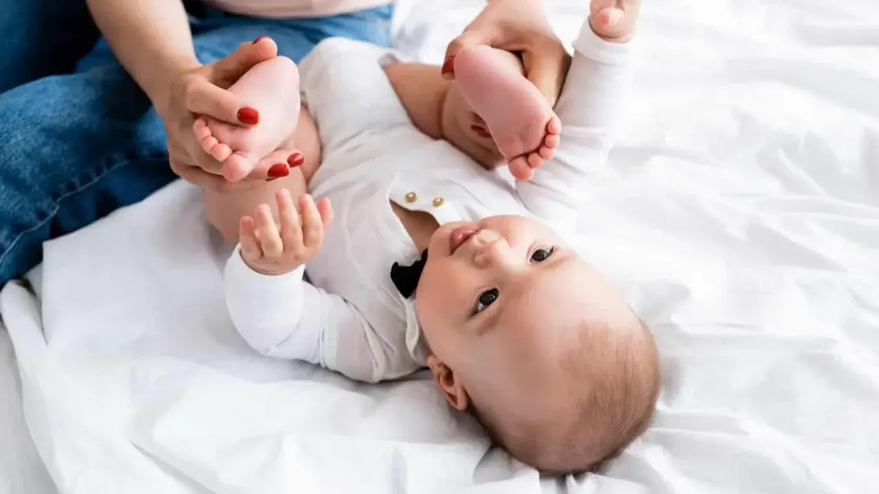 Mama delikatnie ćwiczy nóżki niemowlaka, pomagając mu przy wzmożonym napięciu mięśniowym. Dziecko leży na białym tle.