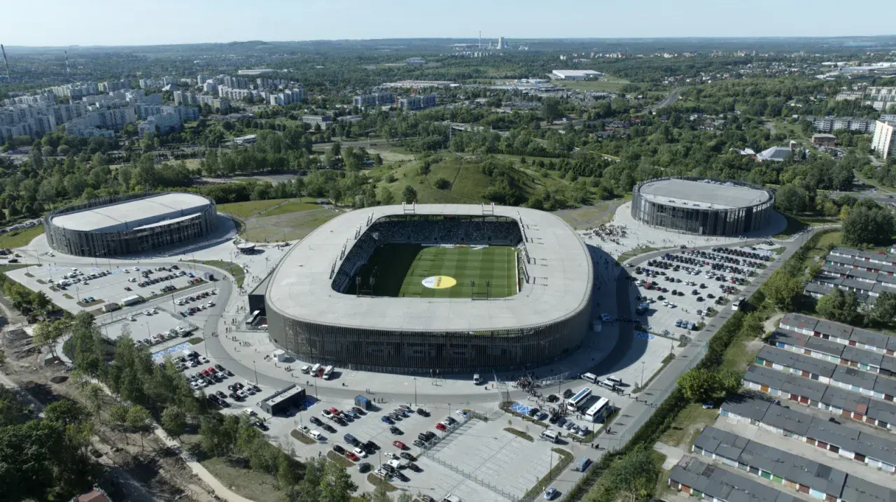 Widok z lotu ptaka na nowoczesny kompleks sportowy, w tym stadion piłkarski i dwie hale widowiskowe. To arena Sosnowiec, otoczona zielenią i parkingami.