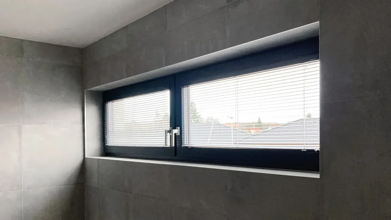Nowoczesne okno z ciemną ramą i żaluzjami, zamontowane w szarej, kafelkowej ścianie. Widok na dachy domów.