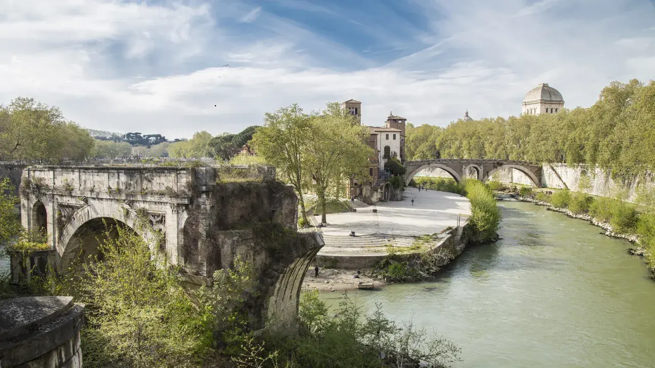 Un ponte rotto, con archi antichi e vegetazione rigogliosa, domina un fiume serpeggiante a Roma.