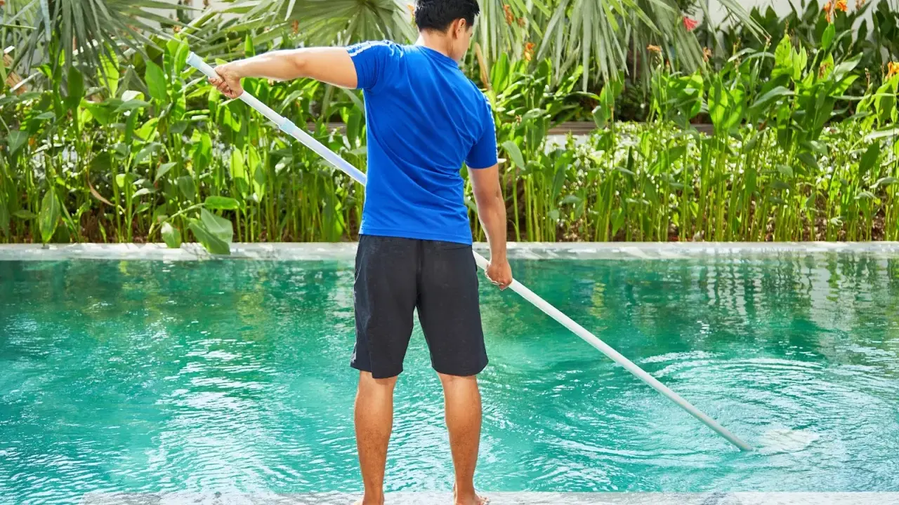 Hombre con camiseta azul y pantalones cortos negros limpia piscina de sal con un limpiafondos.