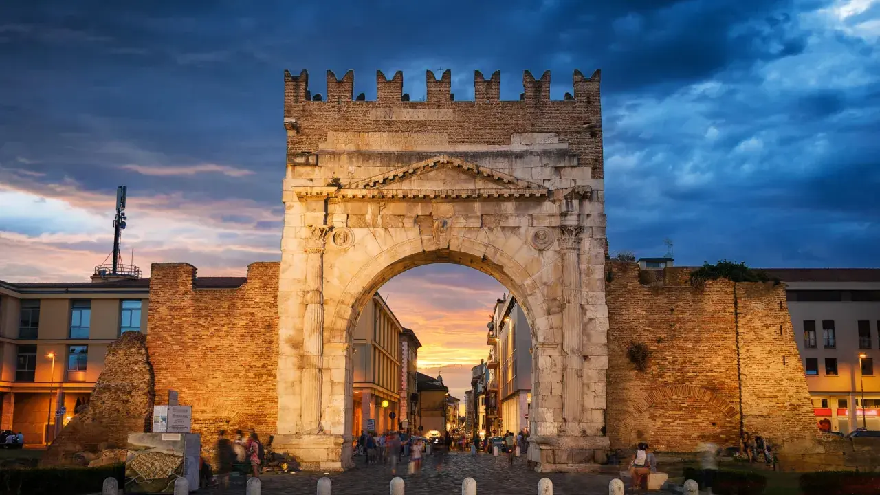 Arco d'Augusto a Rimini al tramonto, con il cielo drammatico e la folla che passeggia. Un'icona della mir fiera Rimini.