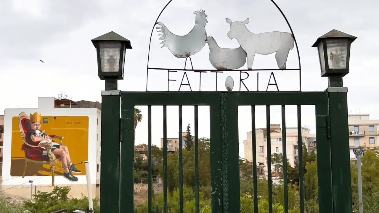 Cancello verde con insegna "FATTORIA" e sagome di animali. A sinistra, murale di un uomo su trono. Danisinni, Palermo.