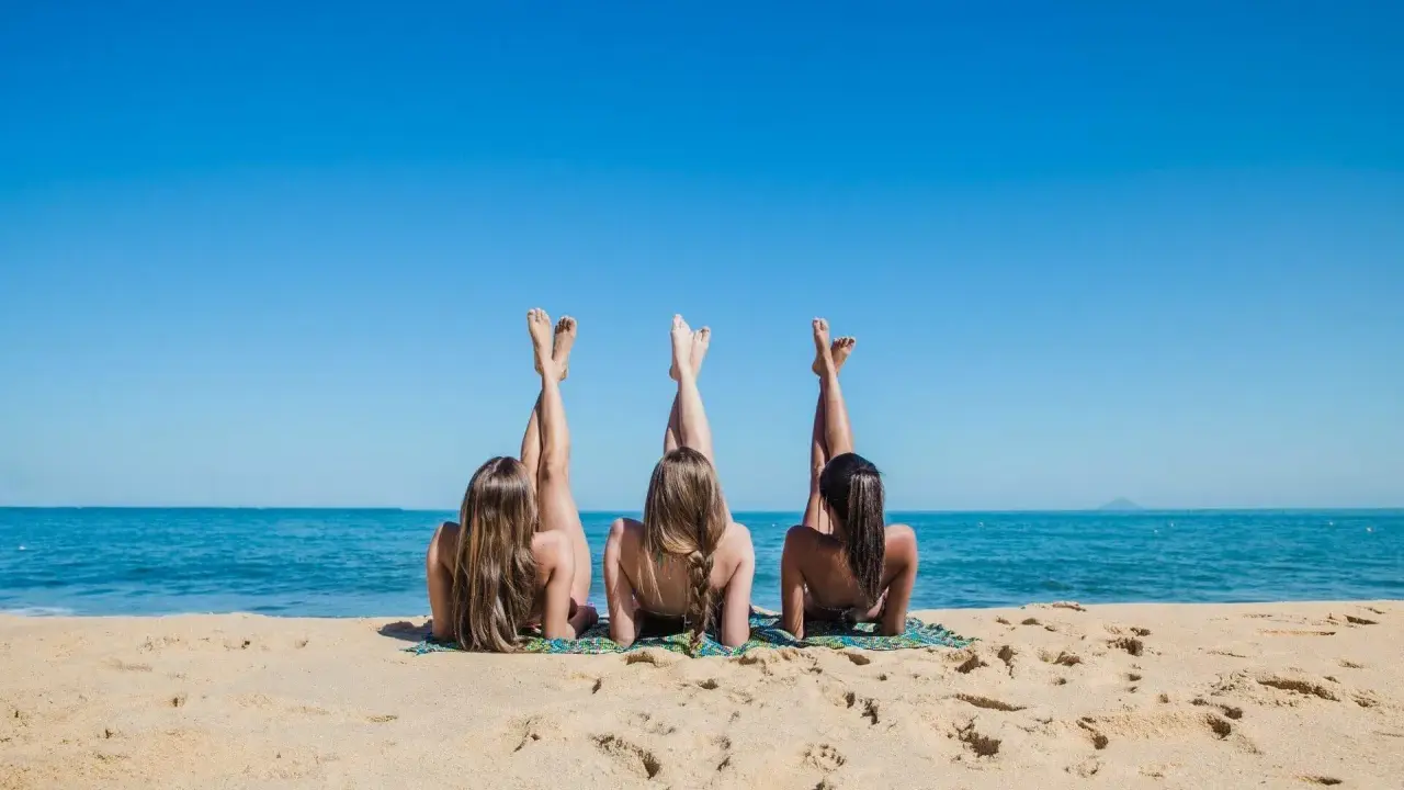 Trzy kobiety leżą na plaży, unosząc nogi do nieba. Słońce grzeje, a morze szumi. Idealny dzień na plaży naturystów.