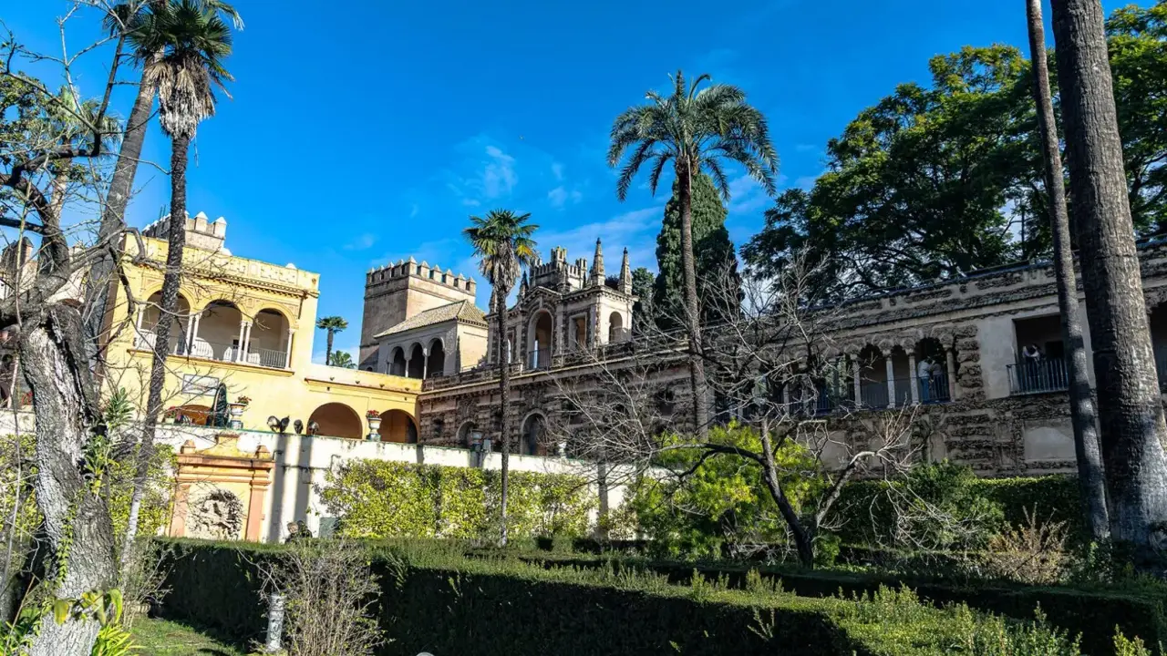 Sonniger Tag im Alcázar von Sevilla. Palmen ragen über kunstvoll verzierte Gebäude und gepflegte Gärten.