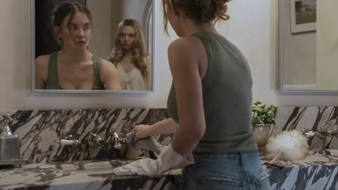 Una ragazza si pulisce il lavandino mentre un'altra la osserva riflessa nello specchio. La scena suggerisce una trama di famiglia complessa.