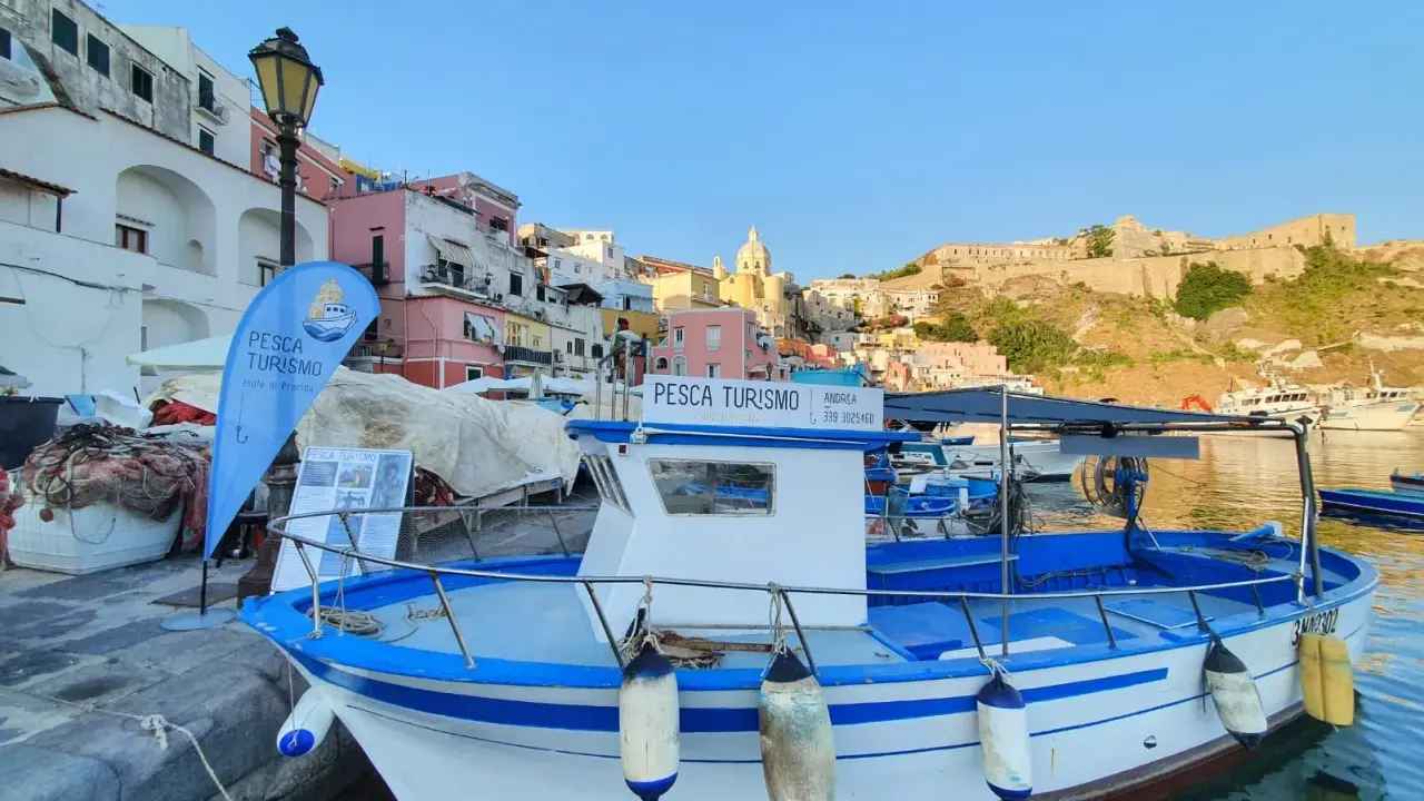 Barca bianca e blu ormeggiata, con bandiera "Pesca Turismo Isola di Procida". Case colorate sullo sfondo.