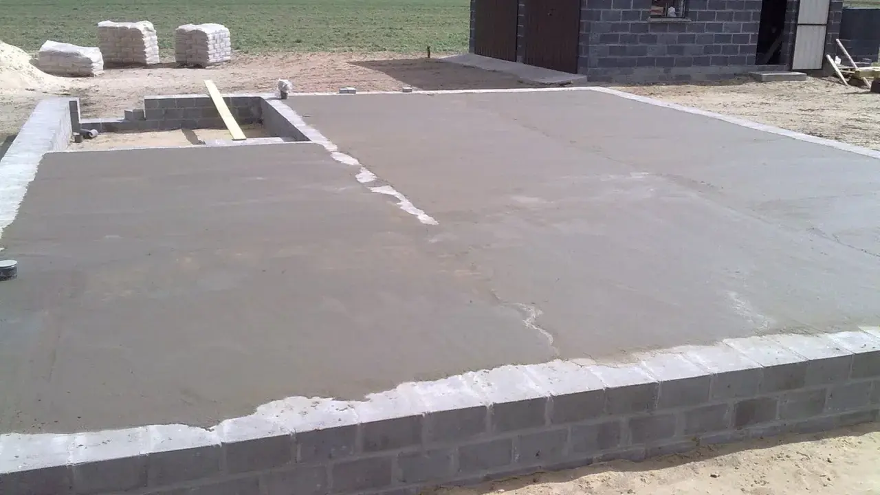 Świeżo wylany beton na chudziaka, tworzący fundament pod budowę. W tle widać materiały budowlane i fragment domu.