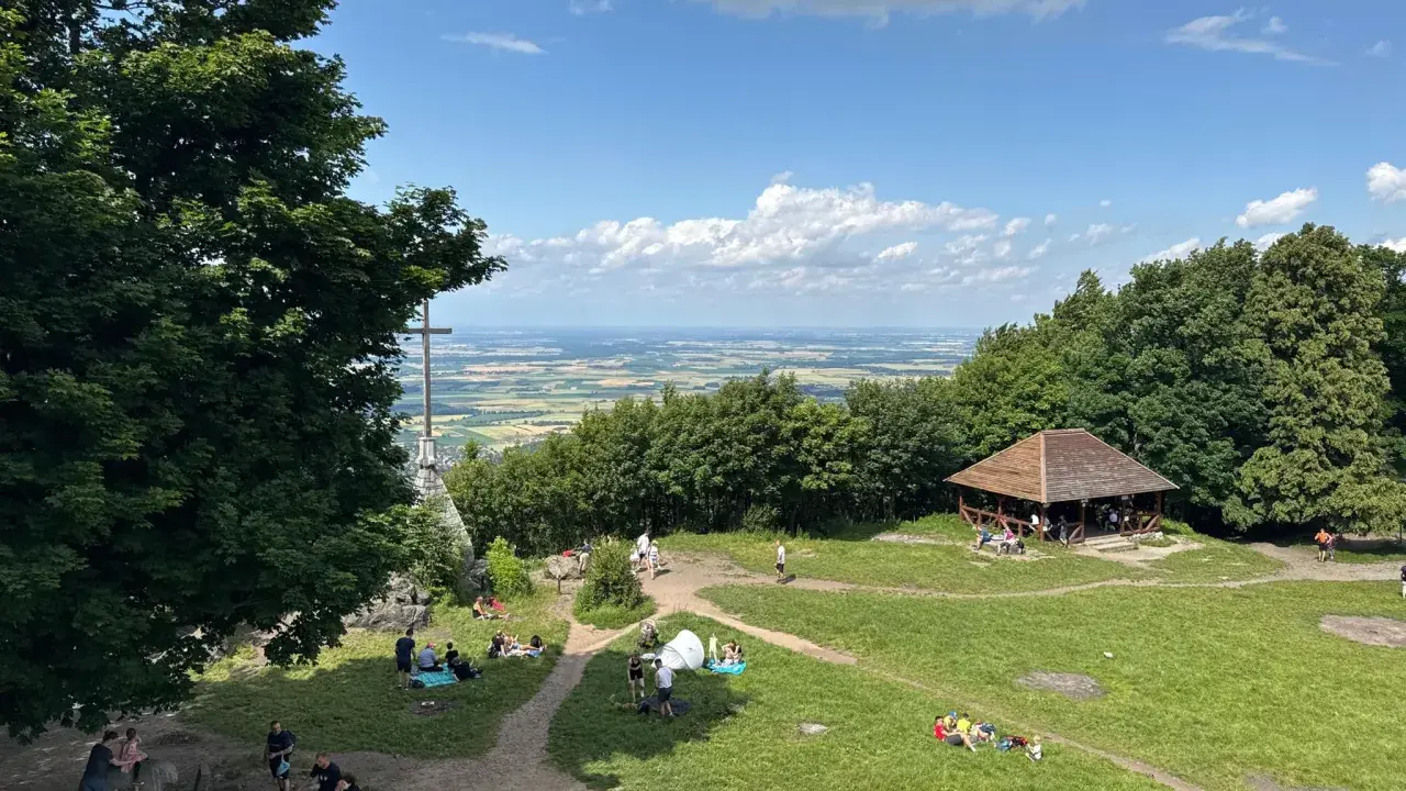 Ludzie odpoczywają na trawie i spacerują po szlakach na górze Ślęża. W oddali widać rozległą panoramę.
