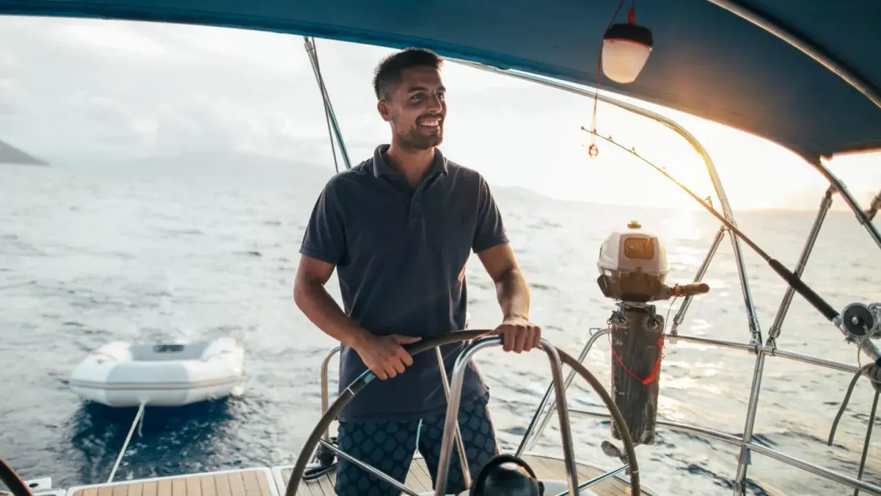 Un homme souriant est au gouvernail, prêt à barrer un bateau. Le soleil brille sur l'eau calme.