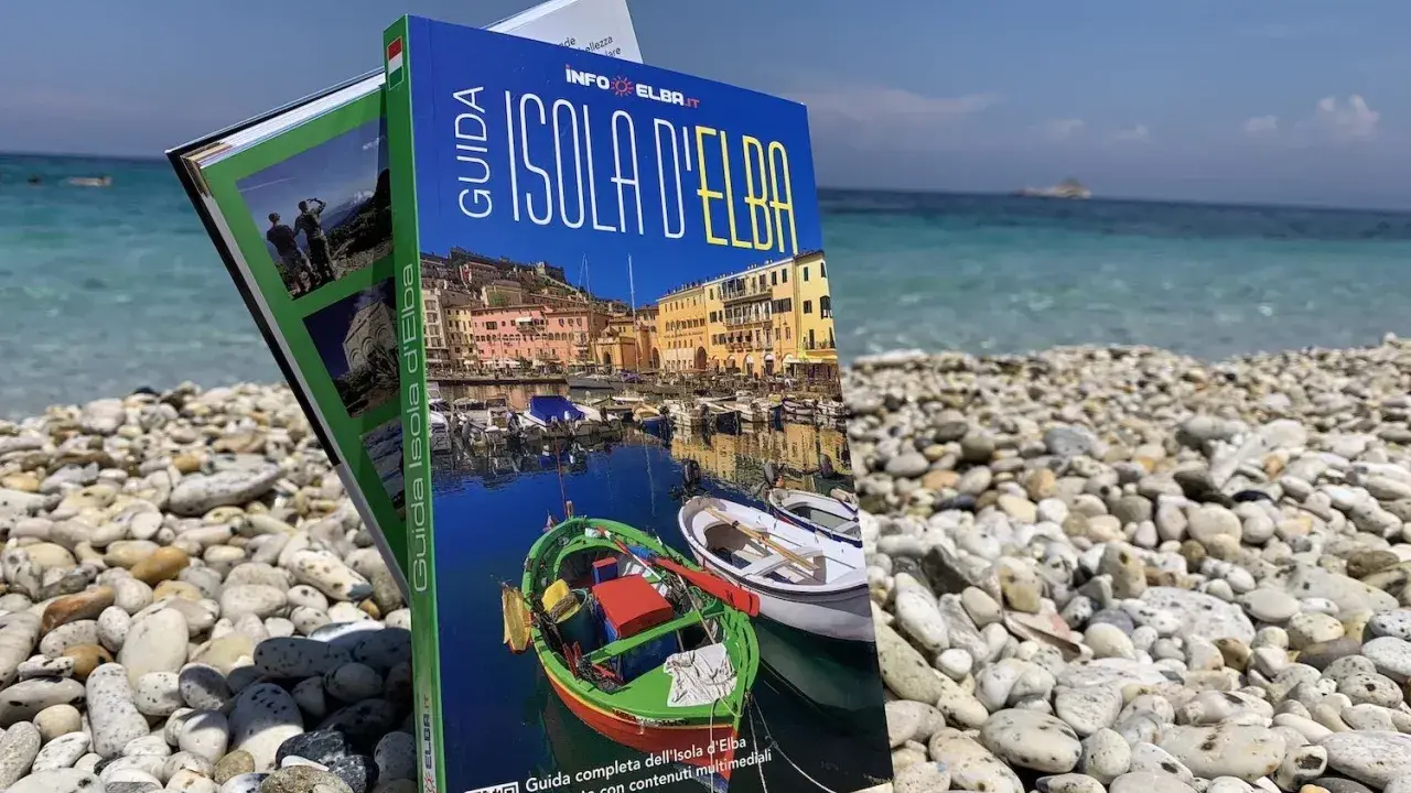 Guida turistica "Isola d'Elba" su una spiaggia di ciottoli, con il mare azzurro sullo sfondo.