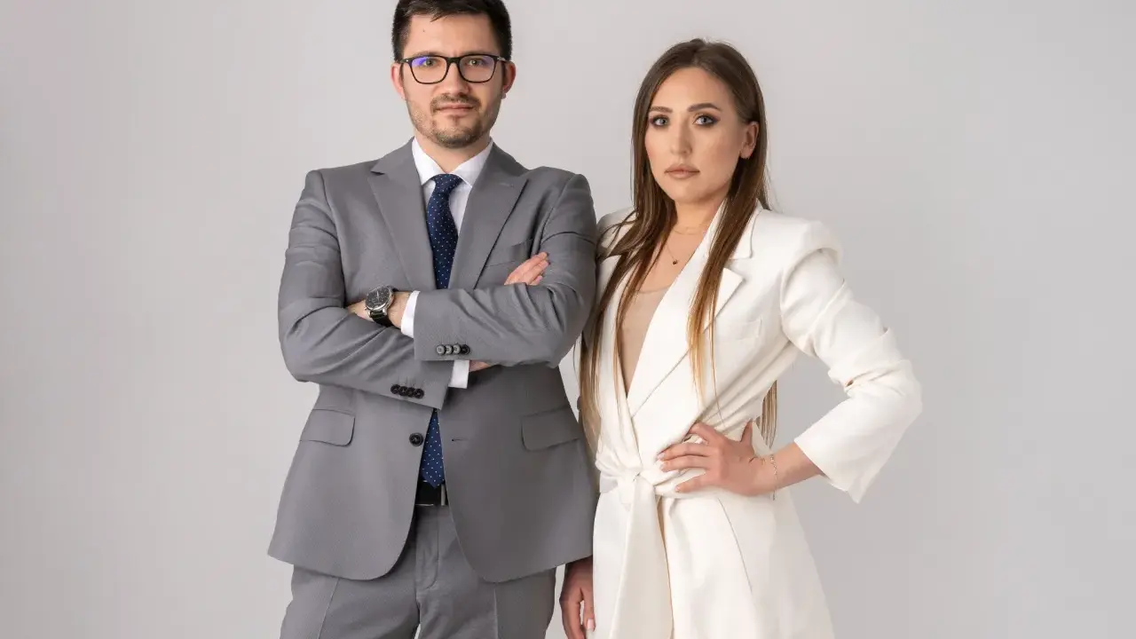 Para prawników w garniturach, mężczyzna w szarym, kobieta w białym, na jasnym tle.