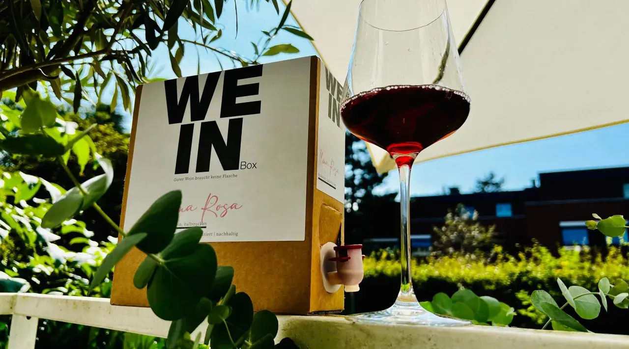 Ein Glas Rotwein steht neben einem Karton mit der Aufschrift "WE IN Box". Der Wein im Karton ist eine praktische Alternative zur Flasche.
