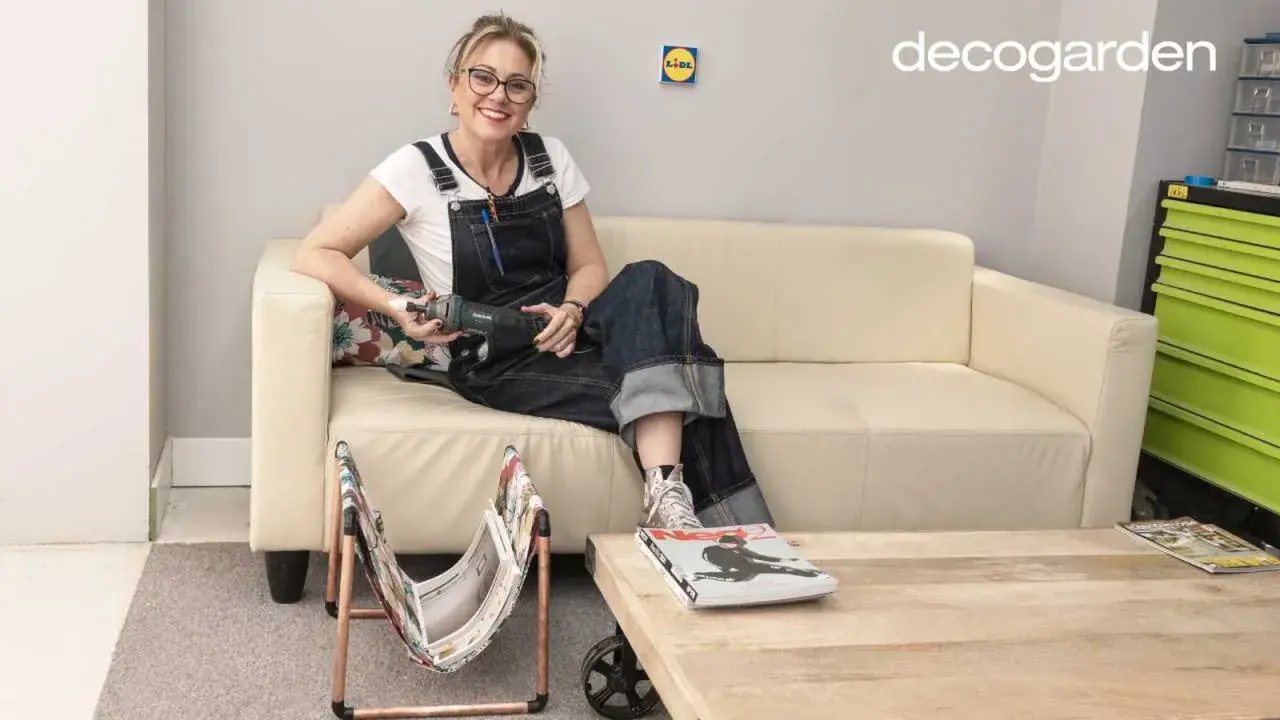 Mujer con overol sentada en sofá beige, sosteniendo una herramienta. A su lado, un revistero diy hecho con cobre y tela.
