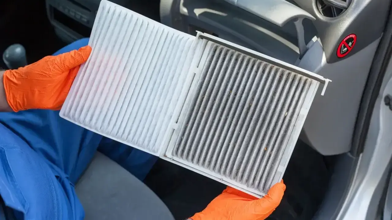 Ręce w pomarańczowych rękawiczkach pokazują brudny filtr kabinowy Toyoty Yaris, gotowy do wymiany.
