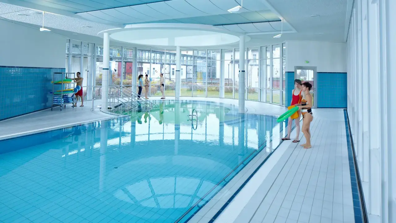 Menschen in Badekleidung am Beckenrand eines Hallenbades mit türkisfarbenem Wasser.
