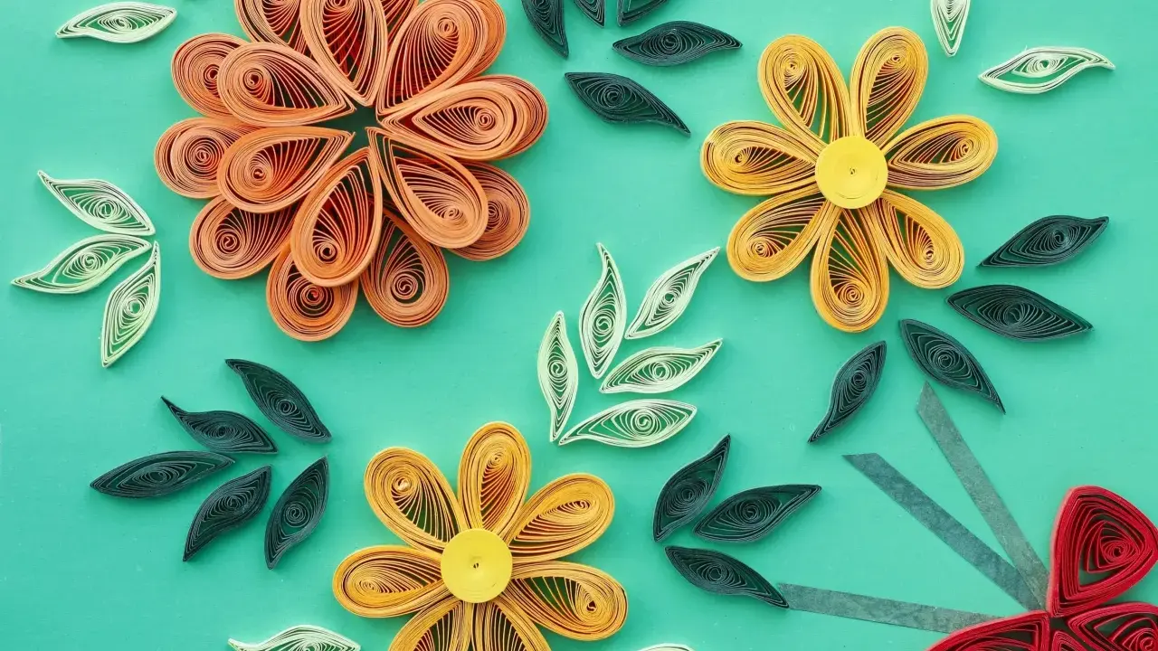 Kwiaty z papieru wykonane techniką quilling dla początkujących. Pomarańczowe, żółte i czerwone kwiaty oraz zielone i białe listki na turkusowym tle.
