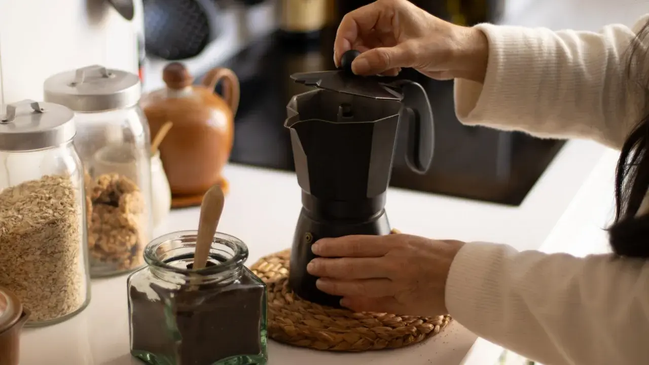 Manos preparando un café con alcohol en cafetera italiana. Hay frascos con avena y café molido.