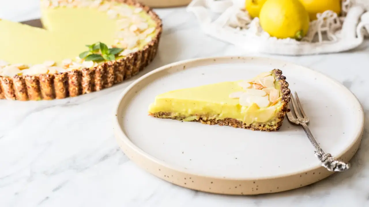 Ein Stück zitronentarte vegan mit Mandelsplittern auf einem Teller. Daneben eine ganze Tarte und frische Zitronen.