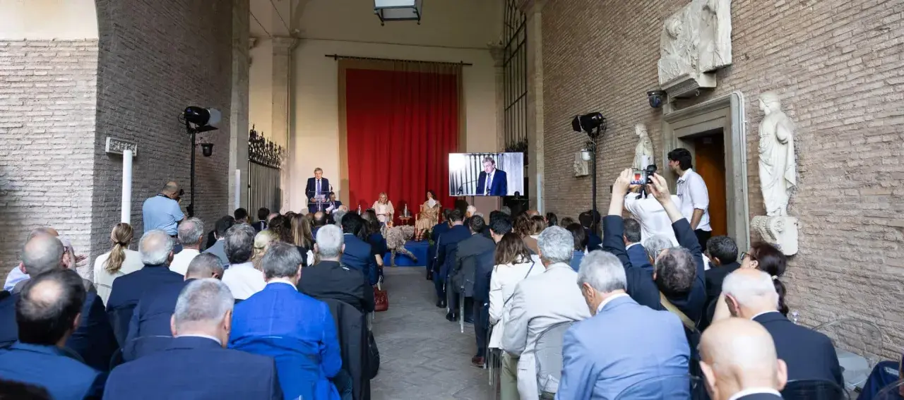 Un evento per il meeting per l'amicizia fra i popoli. Un relatore parla a un pubblico seduto, mentre un altro viene trasmesso su uno schermo.