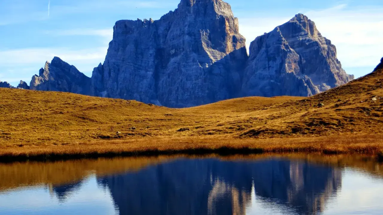 Riflessi maestosi delle Dolomiti nel lago delle Baste, con un prato dorato che separa le acque dalle vette imponenti.