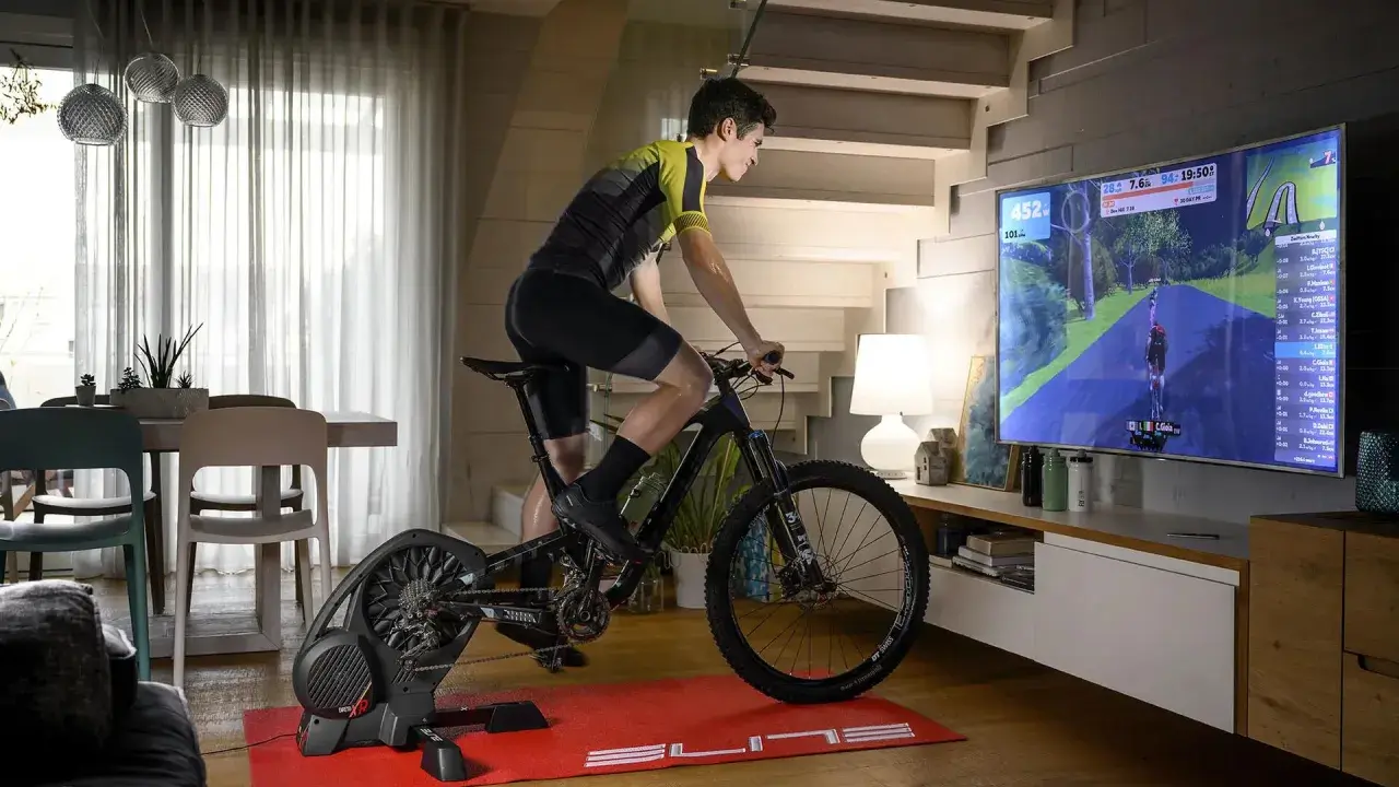 Mężczyzna ćwiczy na rowerze stacjonarnym, korzystając z aplikacji do trenażera, która wyświetla wirtualną trasę na dużym ekranie telewizora.