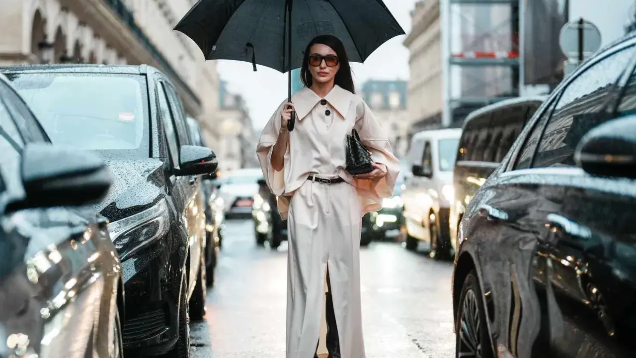 A woman in a chic cream outfit and sunglasses holds an umbrella, showcasing aesthetic rainy day outfits amidst a Parisian street scene with cars.