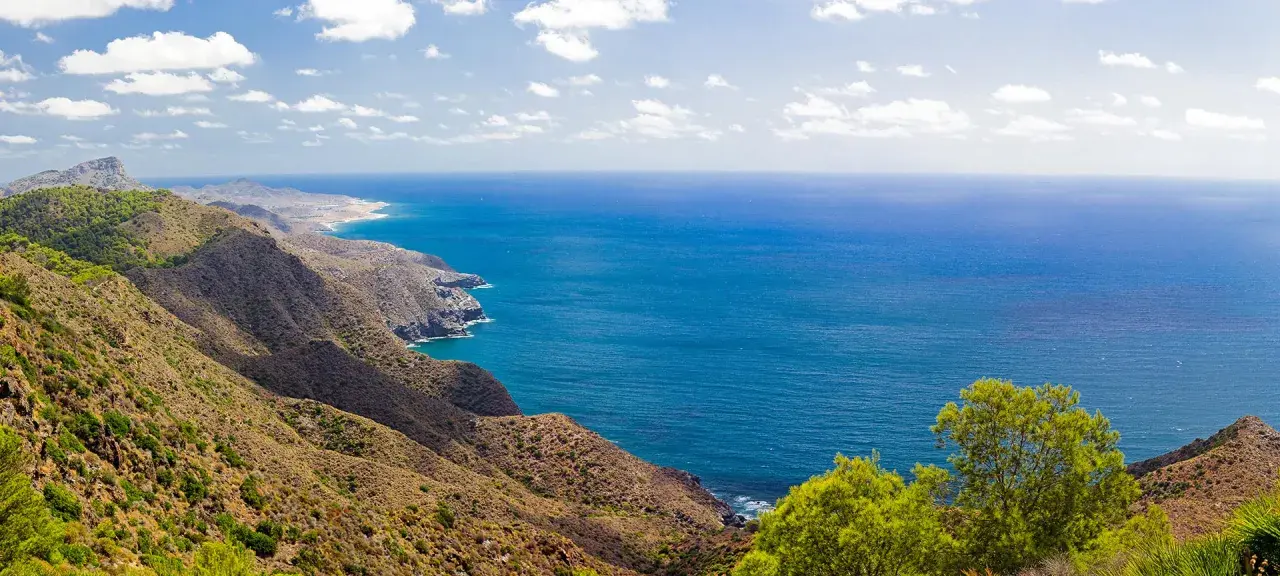 Blick auf die zerklüftete Küste und das tiefblaue Meer bei Cabo de Palos. Grüne Vegetation bedeckt die Hügel, die zum Meer abfallen.