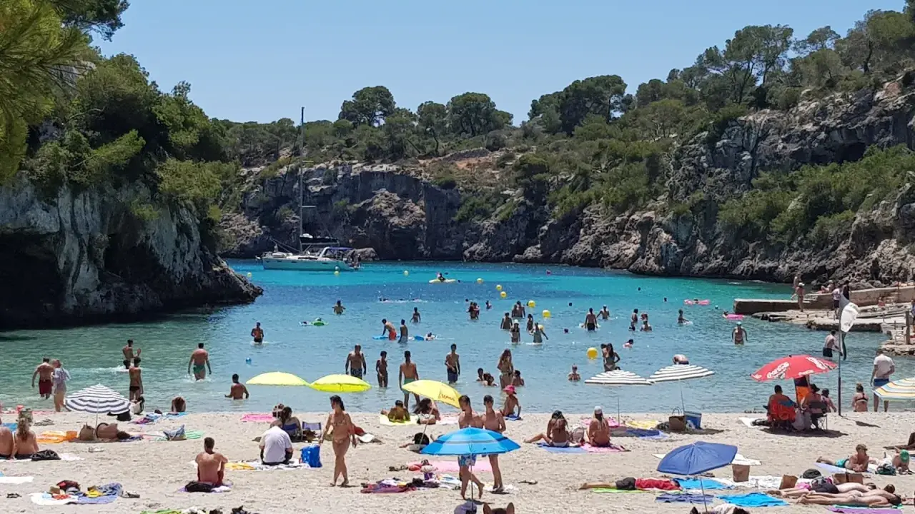 En livlig strand vid Cala Pi, med folk som badar i det turkosa vattnet och solar på sanden under parasoller.