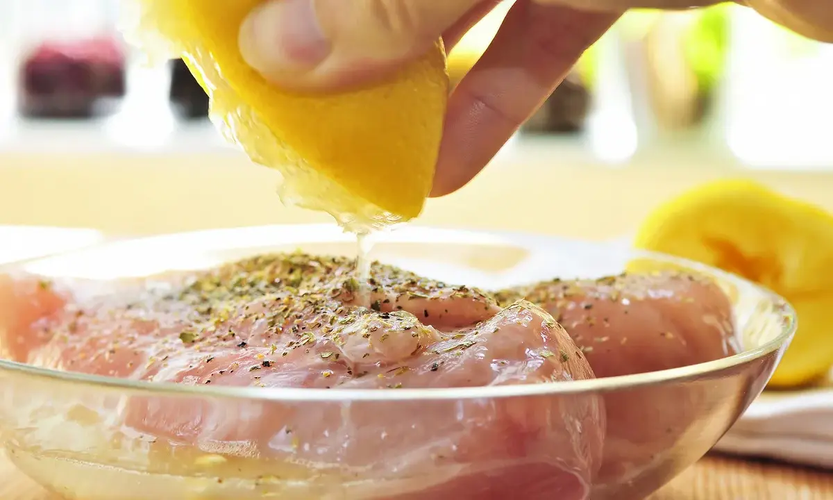 Préparation de la meilleure marinade poulet : un citron est pressé sur des morceaux de poulet assaisonnés dans un bol en verre.