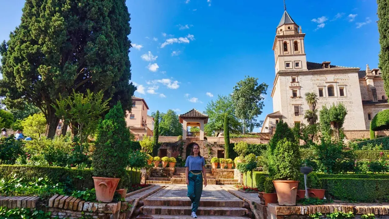 Frau spaziert durch einen üppigen Garten mit Zypressen und einem Turm im Hintergrund. Ein schöner Ort für einen Tagesausflug, vielleicht sogar von Malaga aus.