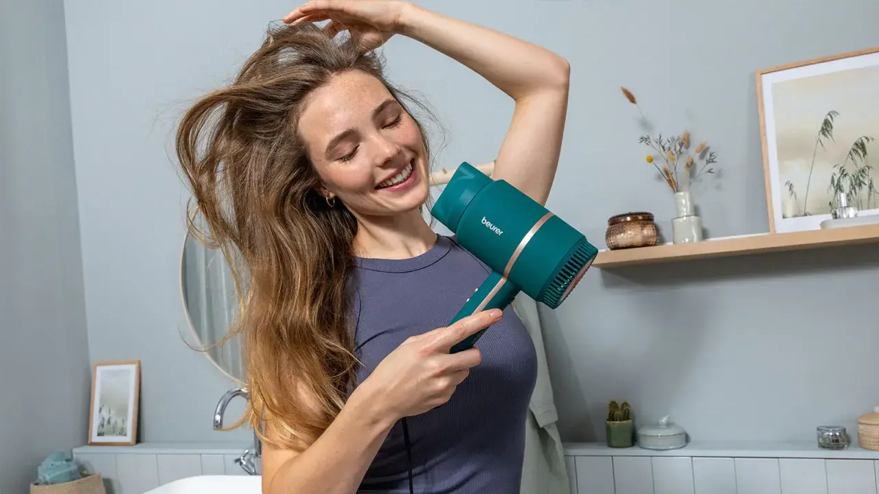 Lächelnde Frau föhnt sich die Haare mit einem türkisfarbenen Föhn. Sie genießt die Pflege ihrer Haare.