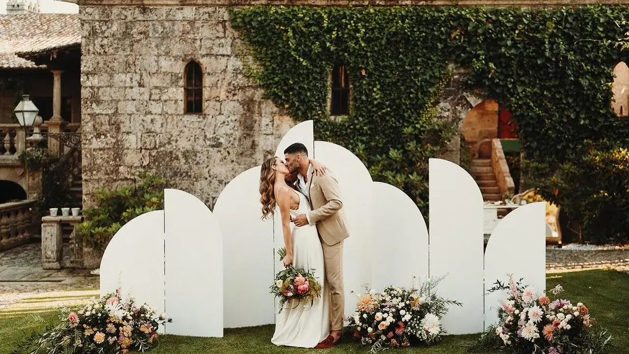 Novios besándose frente a un photocall moderno con arcos blancos y flores. ¡Ideas photocall boda únicas!