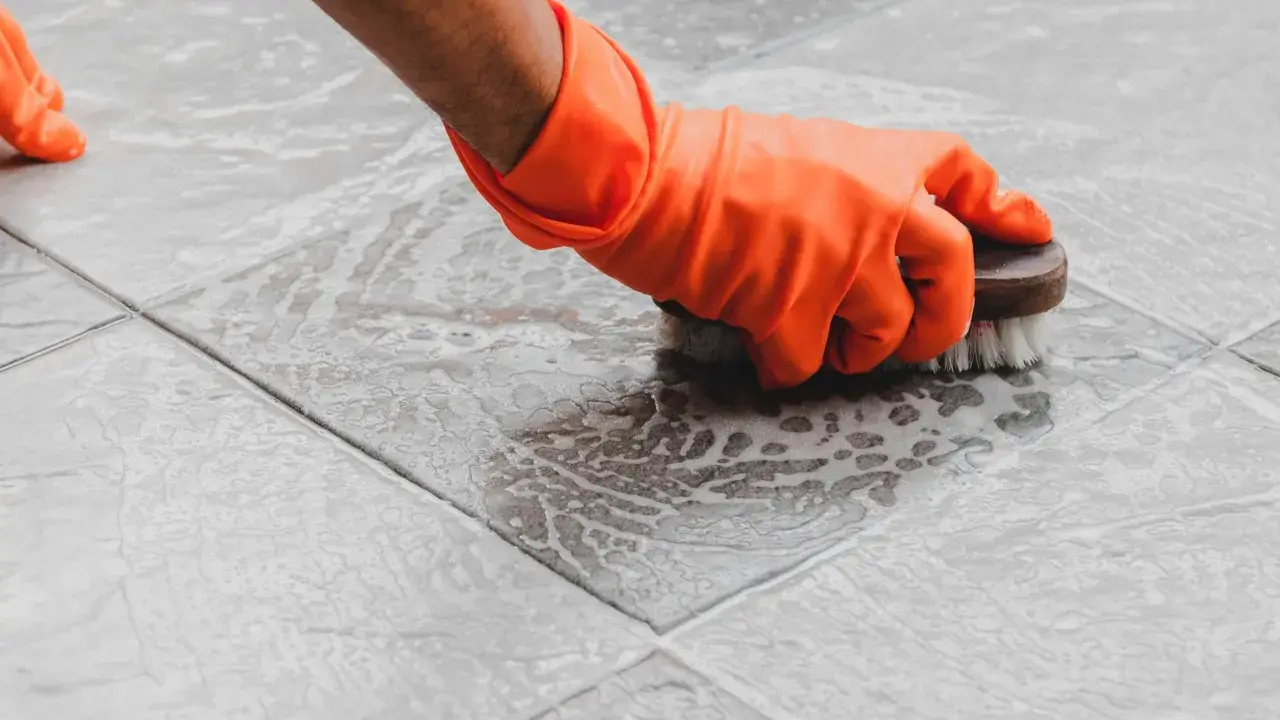 Mano con guante naranja cepillando un suelo de terrazo con espuma. Aprende como limpiar terrazo para que brille.