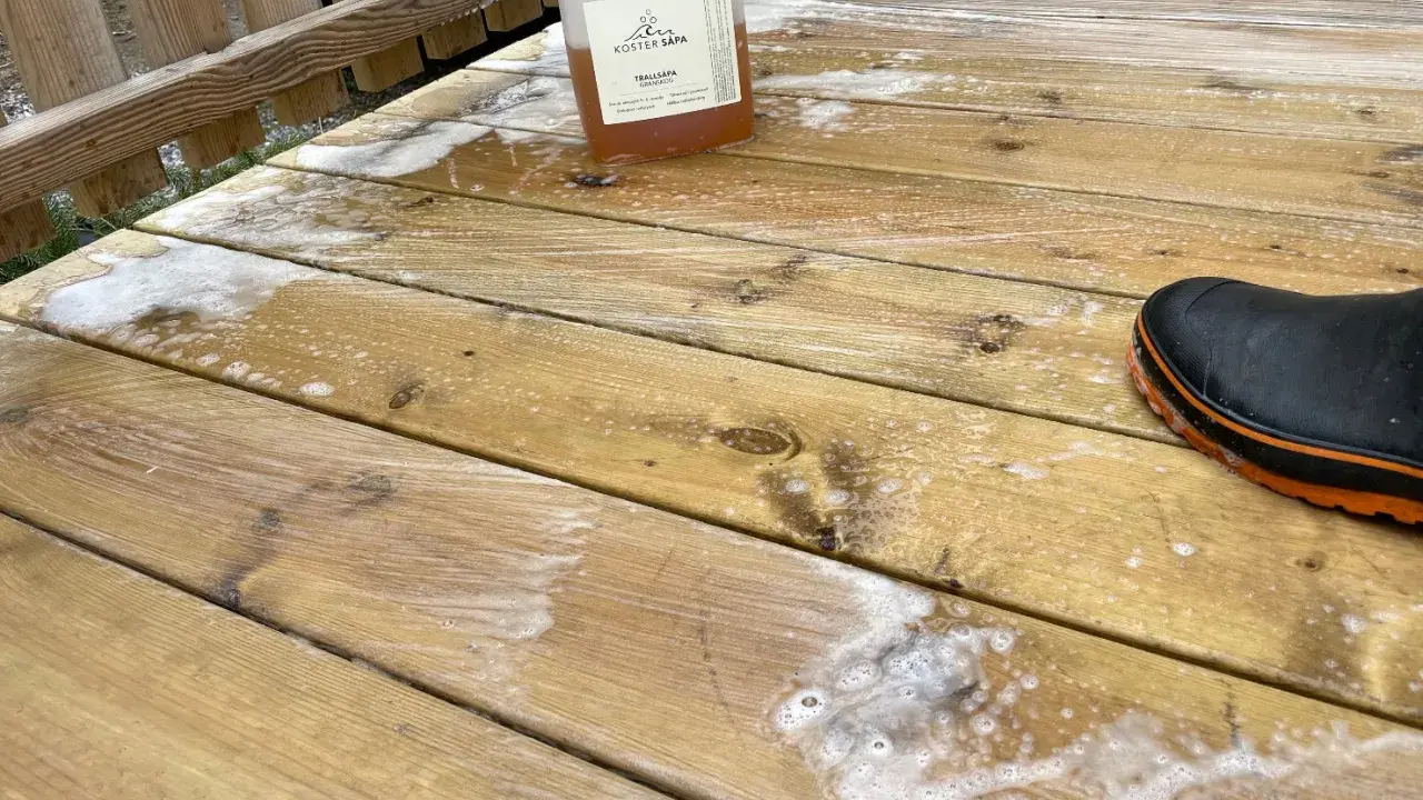 En flaska linoljesåpa står på en altan som håller på att rengöras. En svart stövel med orange kant syns i bildens nedre högra hörn.